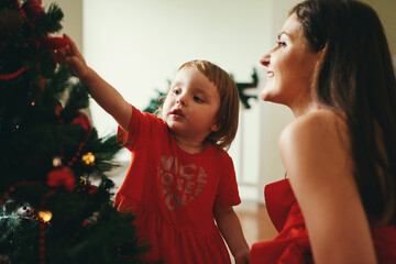 Little girl with mom decorating fir tree with red bell