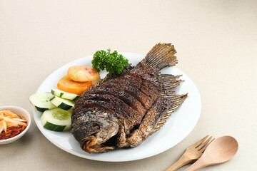 Gurame Bakar Madu, Grilled Gourami with honey and soy sauce. Served with sambal mangga (chilli sauce and grated mango), cucumber and tomatoes in white plate.   
