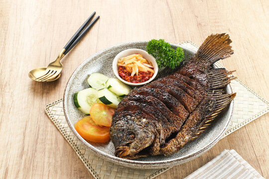 Gurame Bakar Madu, Grilled Gourami with honey and soy sauce. Served with sambal mangga (chilli sauce and grated mango), cucumber and tomatoes in white plate.   
