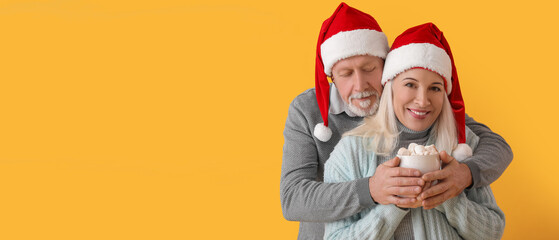 Happy mature couple in Santa hats and with hot chocolate on yellow background with space for text