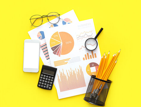Business charts, mobile phone and stationery on yellow background - Powered by Adobe