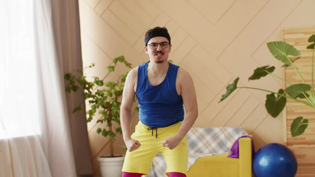 Funny fitness man with glasses, moustache and goatee in bright blue and yellow retro sportswear flexing his muscles talking to the camera. Positive motivation exercise indoors, just do it