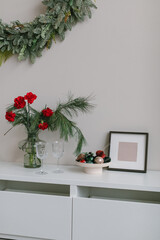 Fototapeta na wymiar Christmas interior decor. Floral composition in stylish vase, Christmas toys in ceramic plate, blank photo frame, glasses on white dresser. Christmas wreath on the wall.