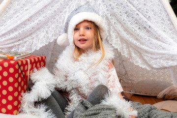 portrait of a cute little girl child wearing a silver color Christmas hat sitting between gift boxes