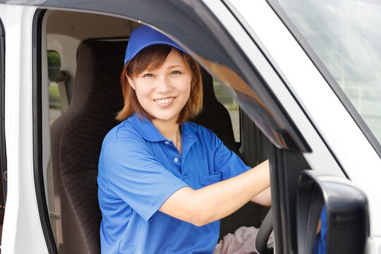 軽トラックを運転する青いキャップとポロシャツを着た笑顔の女性、軽貨物ドライバーとして開業する主婦イメージ。