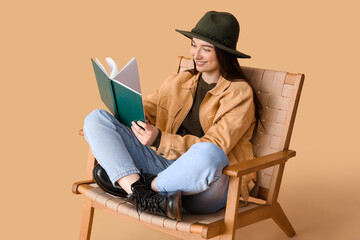 Young woman reading book in armchair on beige background