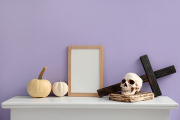 Blank frame with Halloween pumpkins, skull, book and cross on mantelpiece near lilac wall