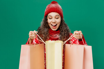 Merry young woman wear red warm cozy knitted sweater hat posing hold package bags with purchases after shopping isolated on plain dark green background Happy New Year 2023 celebration holiday concept