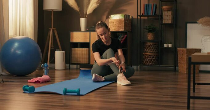 A woman prepares for a workout at home, sits on a mat on the floor and puts on athletic shoes ties the laces. Comfortable footwear.
