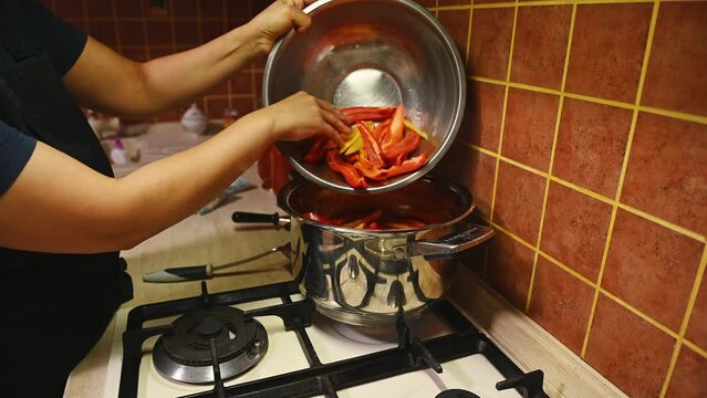 A housewife pours chopped slices of organic ripe homegrown sweet bell pepper into a stream of boiling tomato juice, prepares lecho salad for preservation for the winter.