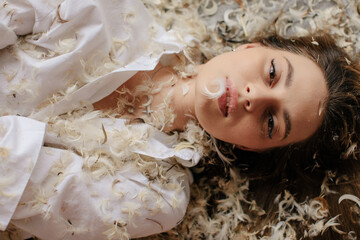 Top view young attractive woman laying on bed covered with feathers and looking at camera - angel  - 537725363