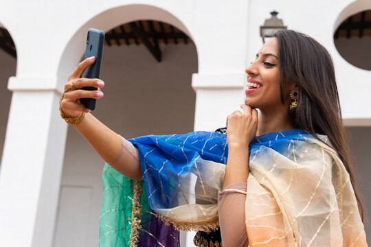 Satisfied Indian woman in traditional outfit taking selfie on mobile phone while standing in garden