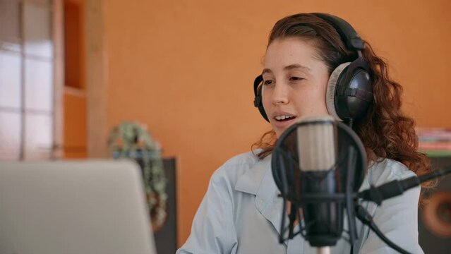 Young podcast show host or content creator talks to laptop with headphones on. In home studio recording session woman is engaged in conversation with audience. Hosting and speaking to fans or callers