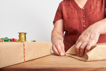 Unrecognizable mature woman wrapping Christmas gifts at home with neutral paper and decorative cords and ribbons. Clean design with copy space.