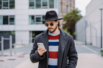 Man in hat and sunglasses using smartphone while walking outdoors in the city. Using mobile phone app.