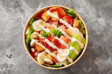 Paper bowl with Caesar salad of lettuce, tomatoes, quail eggs, croutons and smoked salmon on gray stone surface