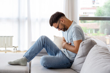 Terrible stomachache. Frustrated handsome young man hugging his belly and keeping eyes closed while lying on the couch at home - obrazy, fototapety, plakaty