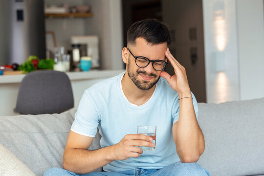 Young man suffering from strong headache or migraine sitting with glass of water on the sofa, millennial guy feeling intoxication and pain touching aching head, morning after hangover concept