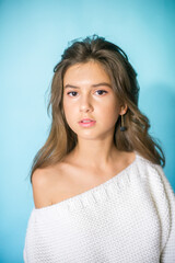 Obraz premium A cute teenage girl with long dark hair in trendy jeans and a white knitted sweater on a blue background in the studio. Fashion for teenagers