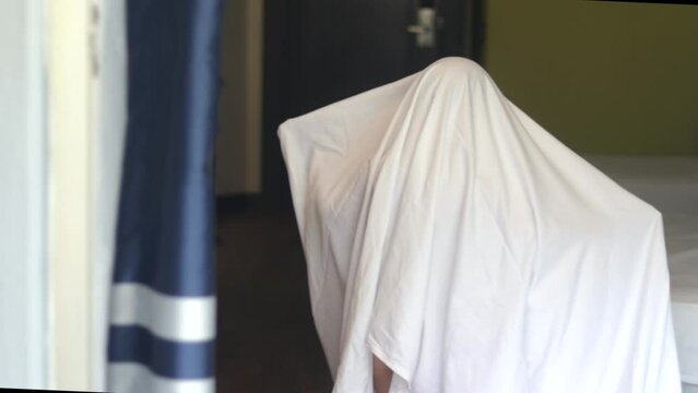 cute kid child boy wrapped in white blanket or covered on head like a ghost jumping moving hands raised up have fun in hotel room bed.halloween trick or treat holiday slow motion 4k