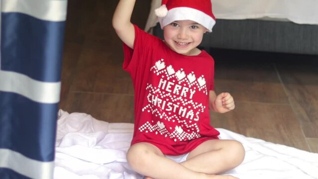 adorable cute little boy sitting on floor on white blanket in red t shirt with merry christmas words and festive ornament elements,santa claus hat cap white pom pom.child kid dancing moving hands sea
