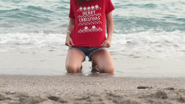 woman in red t shirt with words merry christmas dancing on sands beach against sea waves.happy girl celebrate new year in warm countries vacation travel.female walking from water to coast shell.
