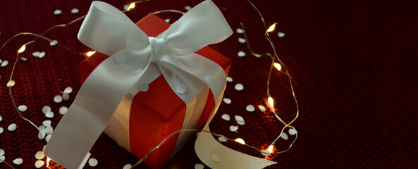 Red gift box with white ribbon on a red knitted background with confetti