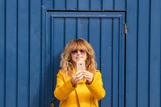 smiling woman with mobile phone in yellow jacket at blue door