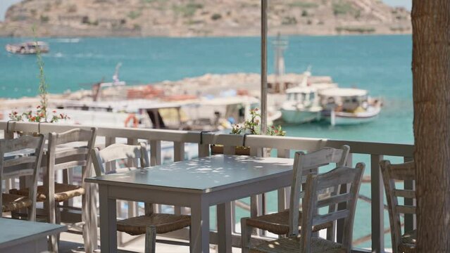 Beautiful and peaceful Taverna in greek island of Crete in sunny Mediterranean. Blue clear water with docks and fishing boats in background. Chairs and tables in traditional greek restaurant