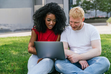 Positive diverse couple sitting on lawn and using laptop