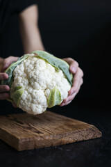 Female hand hold cauliflower over a cutting board on a dark background. Side lighting. Close-up.