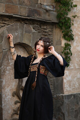 Young woman with long braided hair dressed in ethnic black costume touching wall of old castle. Medieval portrait of aristocratic maiden with jewelry