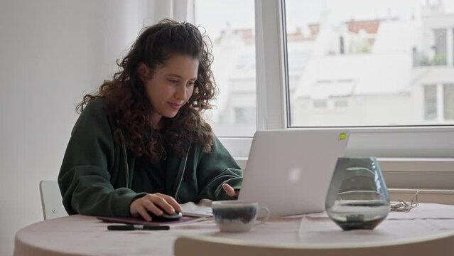 Young woman working from home using laptop computer relaxed and calm. Female dressed casually doing work sitting by the window during a nice day. Enjoys comfort. Works remotely and smiles