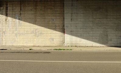 Grunge gray concrete wall of an underpass divided in two by the shadow of the bridge. Cement sidewalk and two lane road in front. Background for copy space