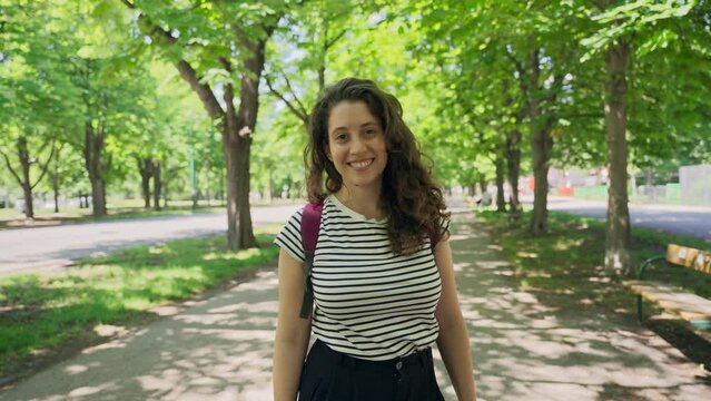 Young woman with backpack smiling looking at camera standing in public park. Portrait of female traveler tourist or student in green park with big trees and bike trails benches and walkway paths