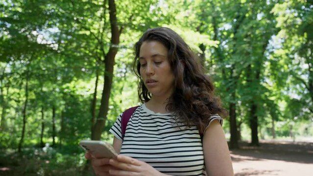 Young woman in forest holding mobile phone looking for location on digital map. Female traveler or tourist in the woods or wilderness searches for signal or whereabouts then finds her path or way