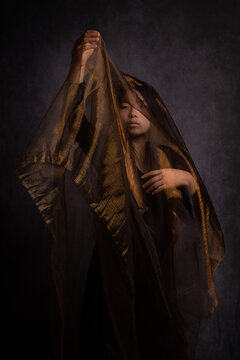 fine art portrait in studio of asian woman in black dress under golden orange veil