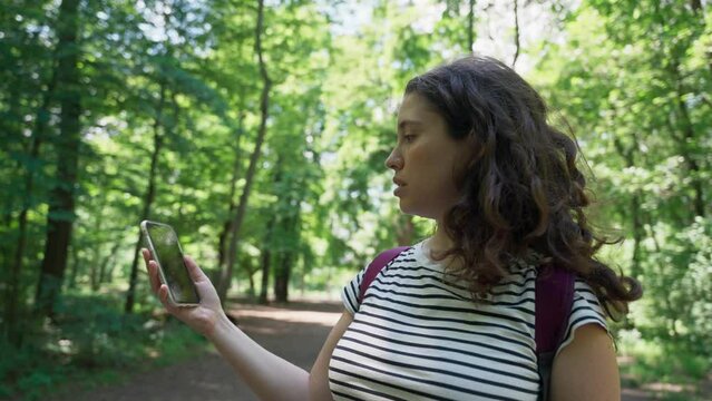 Confused and lost woman with backpack holds phone trying to find her location. Young female standing in forest woods looking for her whereabouts or mobile signal. Unsure where to go or where she is