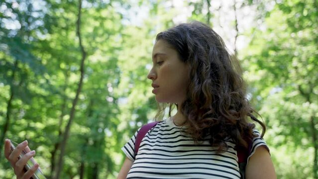 Young woman in forest holds phone and looking for her location on digital map. Female traveler or tourist lost and confused about where she is or has no mobile connection or signal. Looks at a map app