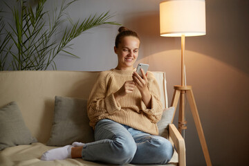 Image of smiling delighted young adult woman wearing casual clothing sitting on sofa and using...