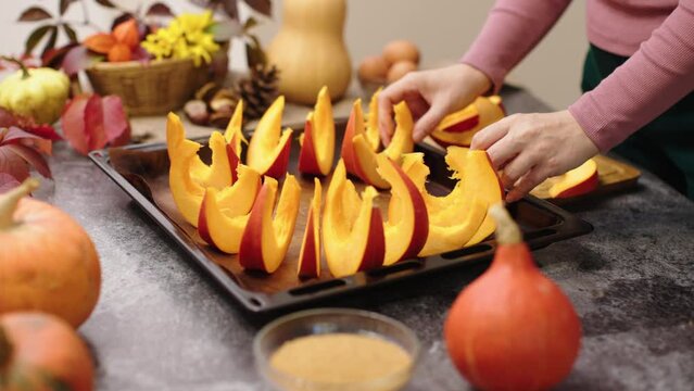 hands put sliced orange pumpkin pieces on baking pan prepare baked in oven baking raw pumpkin sweet dessert traditional autumn food thanksgiving holiday decoration on kitchen table