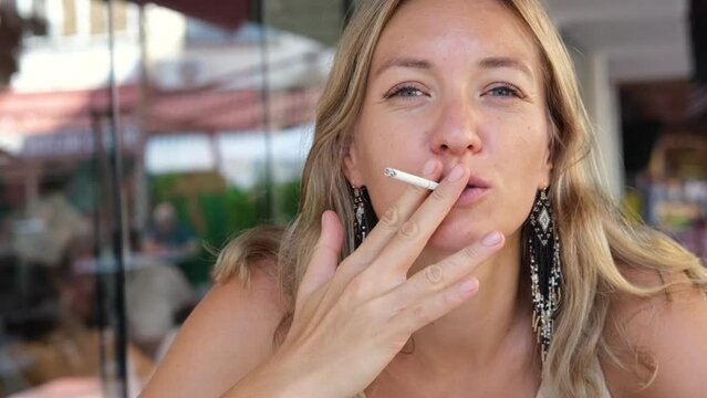 Beautiful blond woman smoking a cigarette outside in public place. Unhealthy lifestyle.