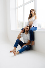 Young happy pregnant couple in love barefoot sitting on windowsill. Laughing and smiling man and woman. Happy pregnancy concept