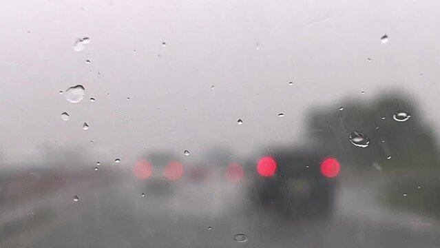 Close up of rainy depressive weather over windscreen and red blurred lights of car traffic in background with wiper cleaning windshield from raindrops