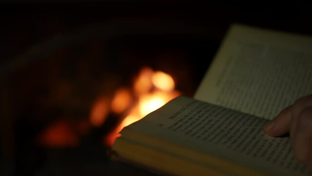 A man reading a book close to an open fire in a fireplace in a dark room