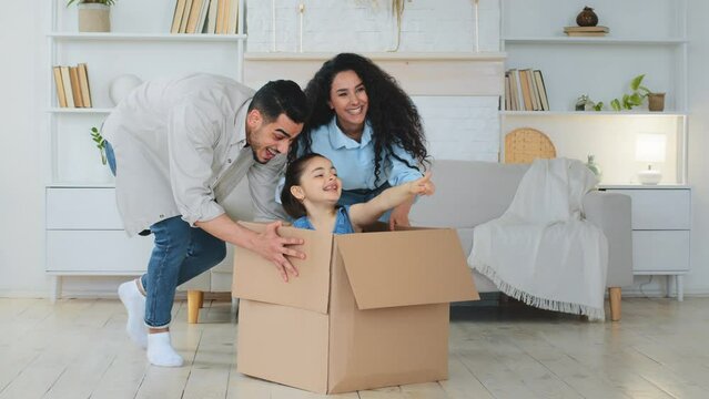 Multiracial multiethnic mother and father Indian Hispanic Caucasian parents push cardboard boxes with little cute child girl daughter ride inside homeowners family playing fun in new house relocation