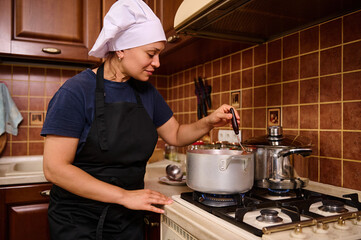 Multiethnic woman in white chef's cap and black apron, stands by the stove, stirring boiling tomato...
