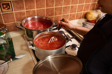 Rear view of a woman in white chef's cap and black apron, stands by the stove, stirring boiling tomato juice while preparing homemade passata from ripe organic juicy tomatoes in the kitchen at home