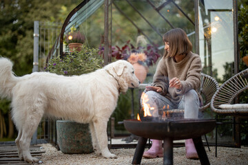 Young woman cares her dog while spending evening time at cozy atmosphere by the fire in garden