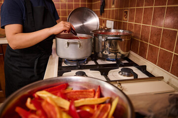 Close-up. Cropped view of a housewife using ladle, stirring the boiling tomato juice in a saucepan, preparing salad lecho with organic tomatoes and peppers. Canning sweet bell pepper for winter
